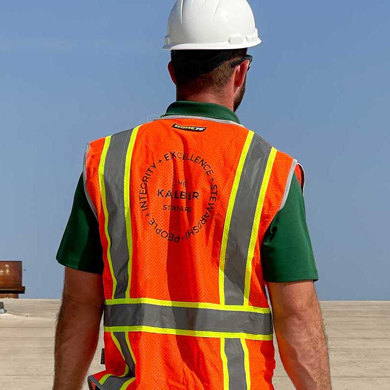 roofer with white helmet orange vest with The Kaliber Standard printed on it
